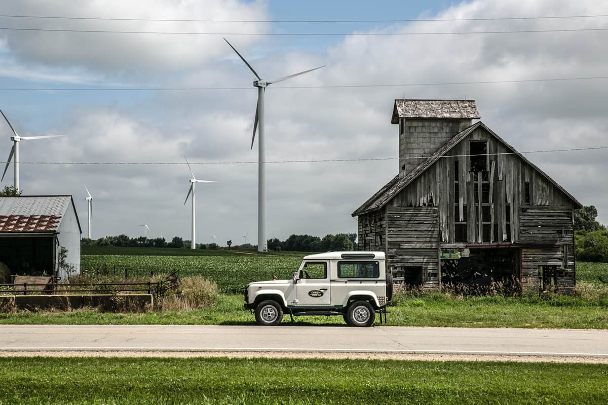 A white Land Rover Defender 90 parked beside a weathered Midwestern barn with wind turbines rising over a summer cornfield