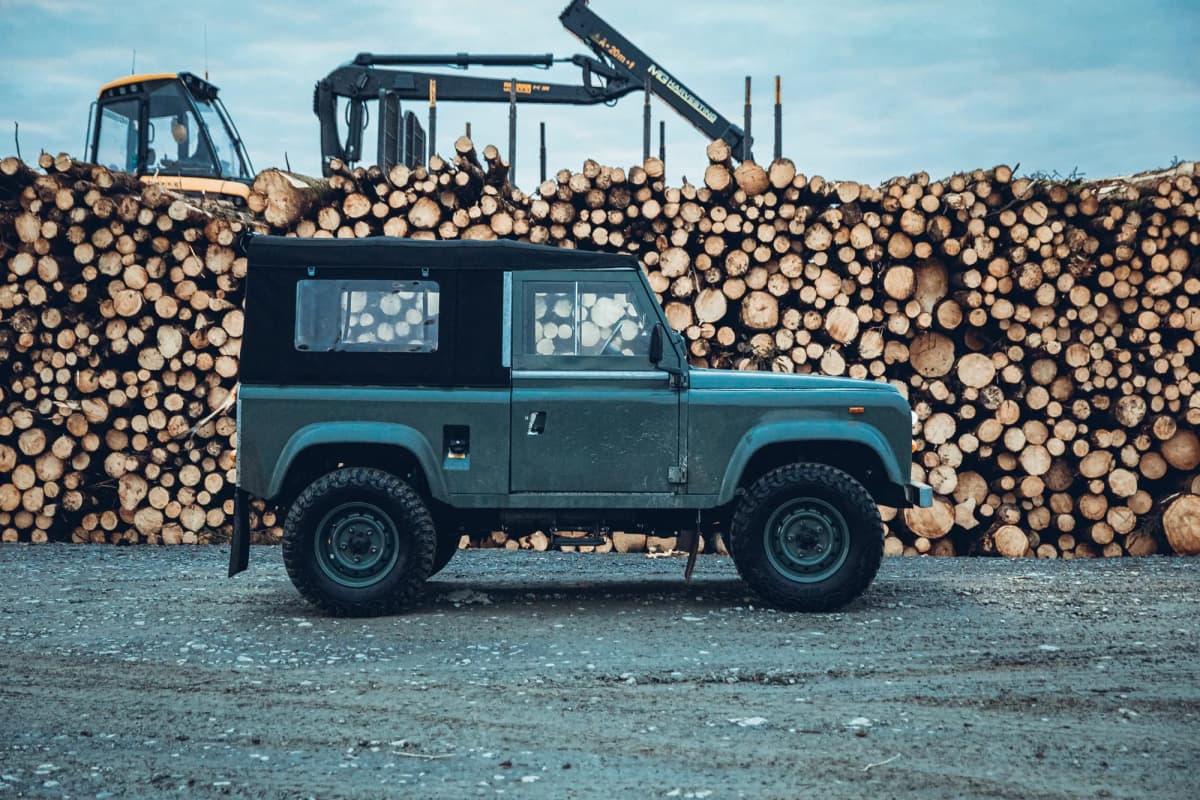 A blue Land Rover Defender 90 parked in front of a stacked log yard at first light