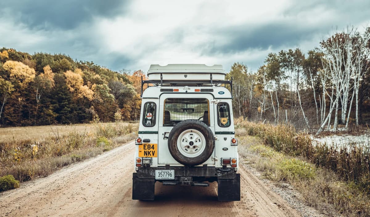 A white Defender 110 heading down a Minnesota dirt road in autumn, kit boxes on the roof